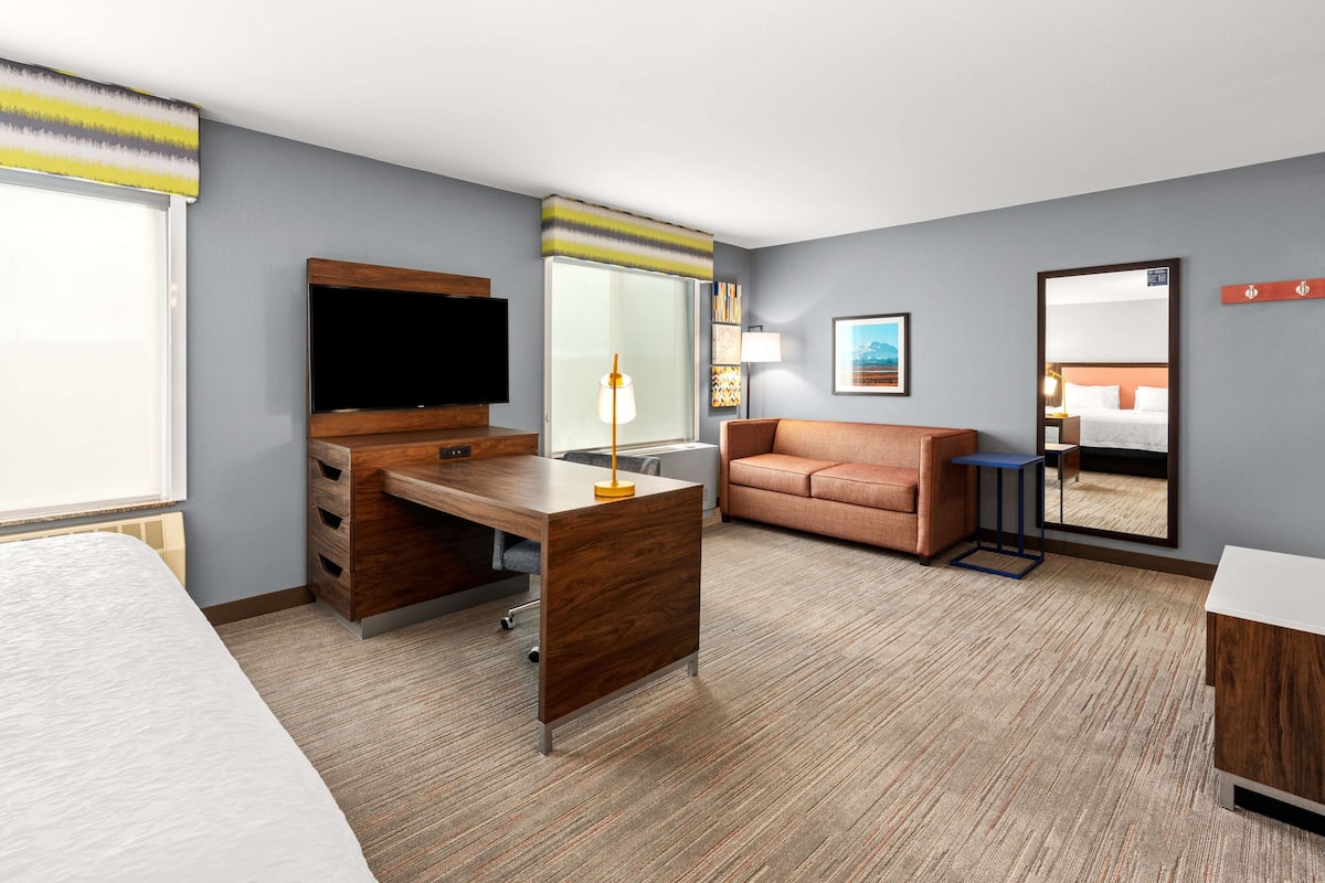 A hotel room with a bed, a desk with a TV, a sofa, a mirror, and a window with blinds.