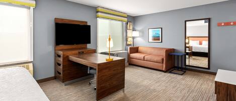 A hotel room with a bed, a desk with a TV, a sofa, a mirror, and a window with blinds.