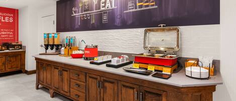 A buffet setup with various food items and containers, a large wall with kitchen utensils and the word "EAT" prominently displayed, and a wooden cabinet with drawers.