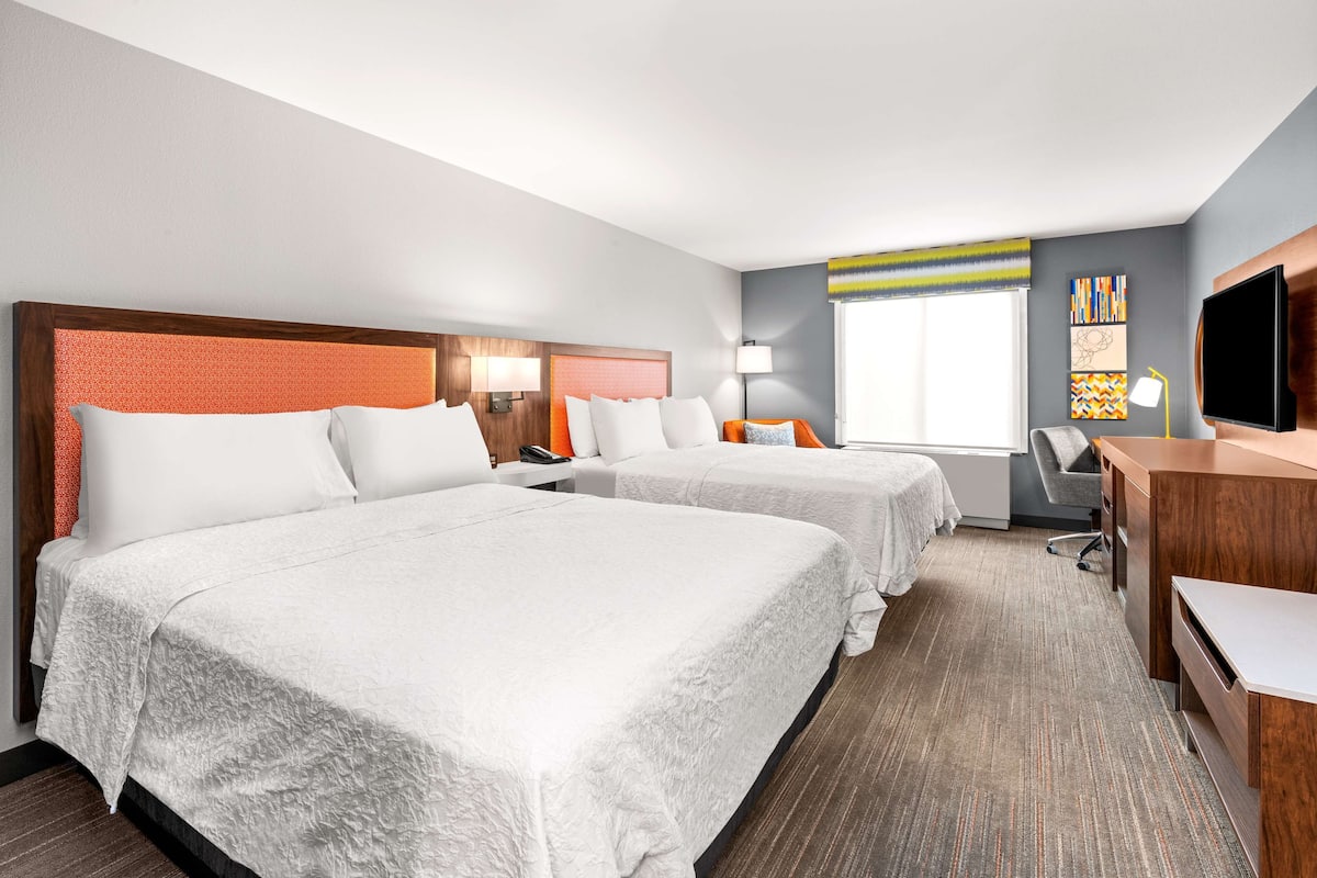 A hotel room with two beds, a desk, a chair, a TV, and artwork on the wall.