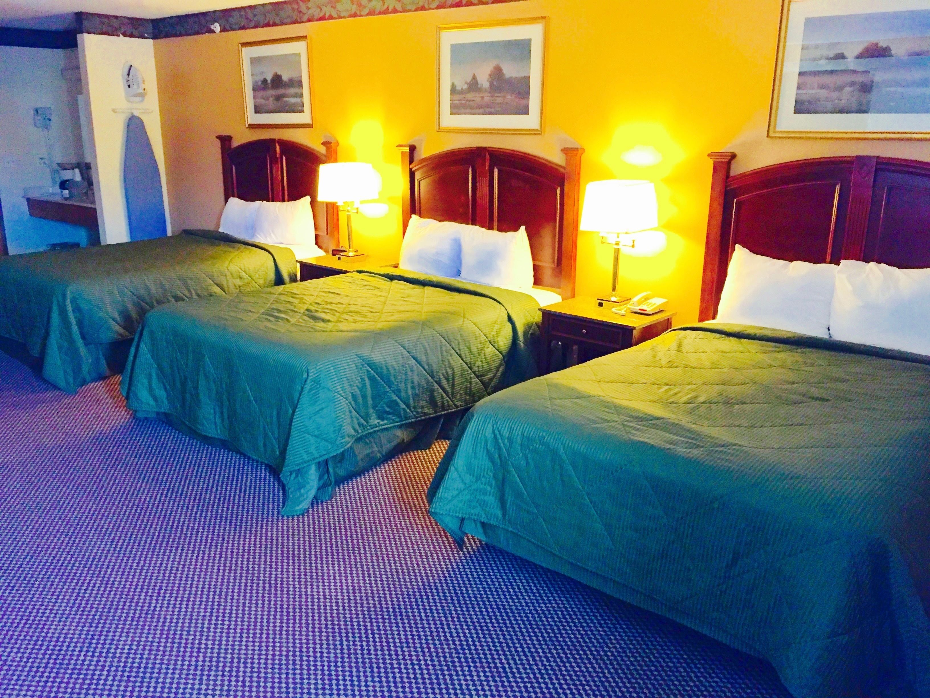 A hotel room with three single beds, each with a green bedspread and white pillows. There are two bedside tables with lamps, and the walls are painted yellow.