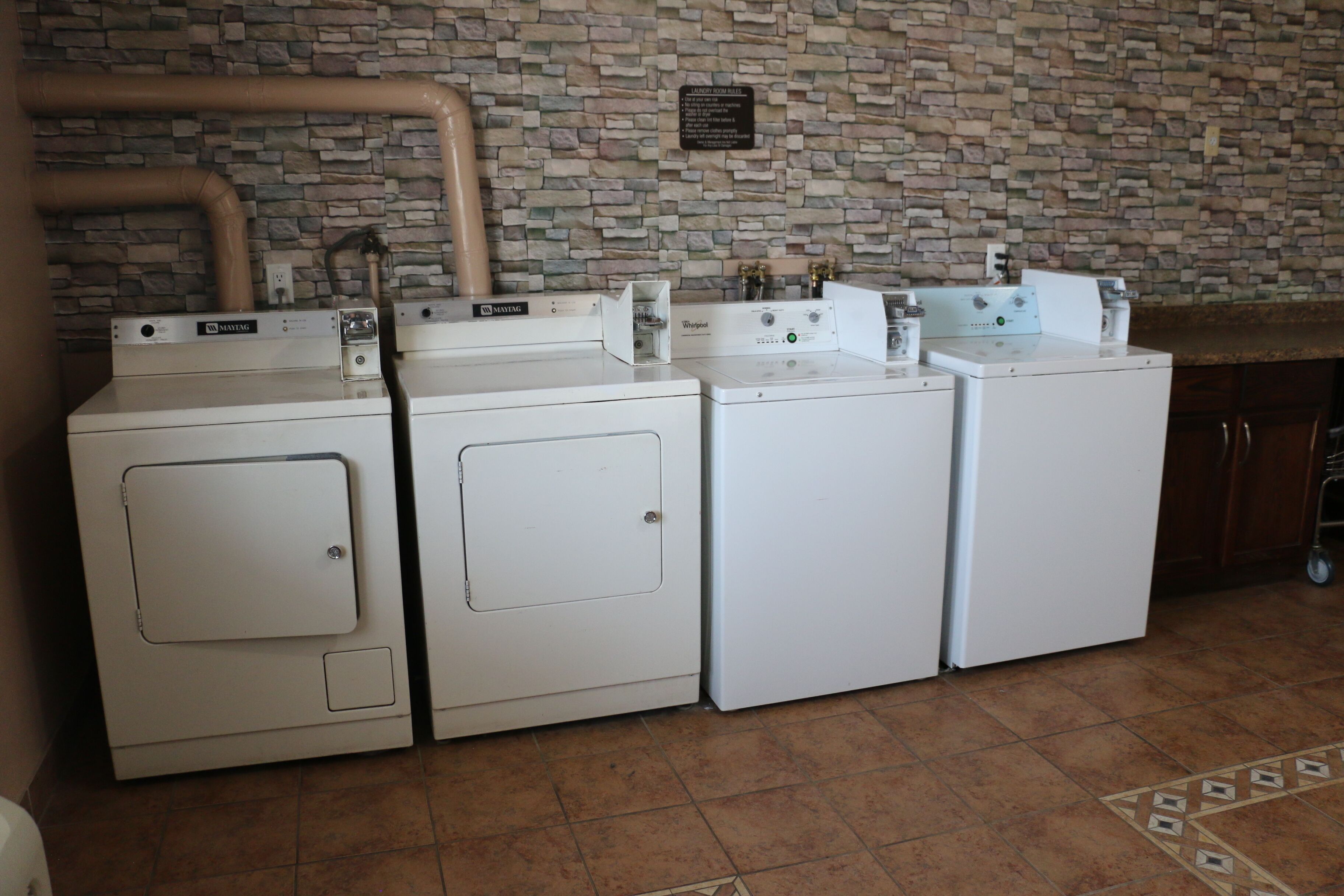 A laundry area with four white washing machines, a stone-patterned wall, and a tiled floor.