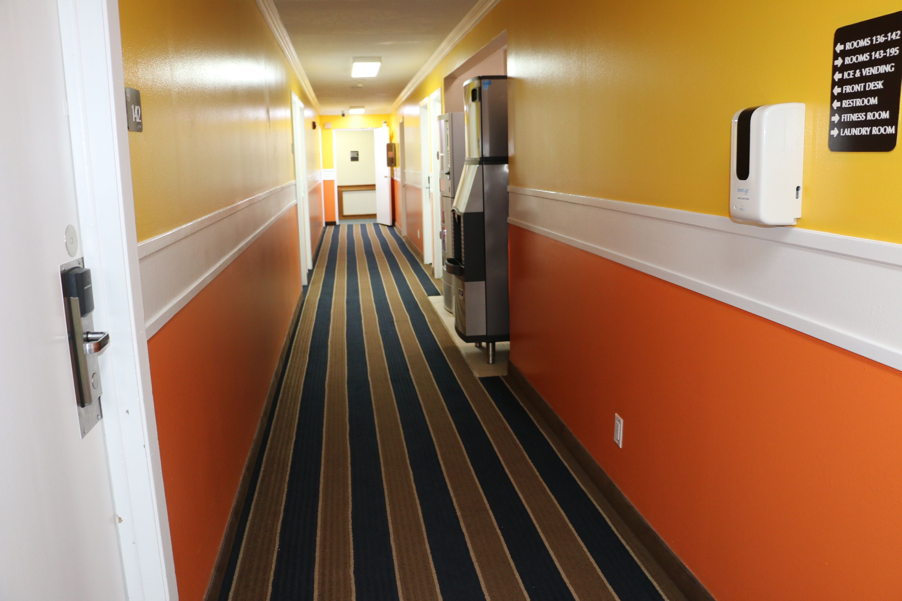 A hotel corridor with a striped carpet, a vending machine, and a sign listing amenities like rooms, ice and vending, front desk, restroom, fitness room, and laundry room.