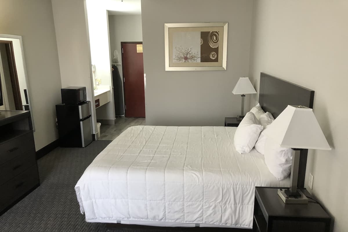 A hotel room with a large bed, a nightstand with a lamp, a dresser, a microwave, and a door leading to a bathroom.