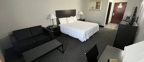 Une chambre d’hôtel avec un lit, un canapé, un bureau, une chaise, une table de chevet, une lampe, un tableau au mur et une porte.