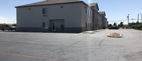 A large, single-story building with a flat roof and a grey facade. There are multiple windows on the front and side of the building. The parking lot in front of the building is empty, with a few cars parked in the distance.