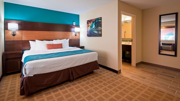 Habitación de hotel con cama, mesitas de noche, cabecera, un cuadro y baño visible a través de una puerta abierta.