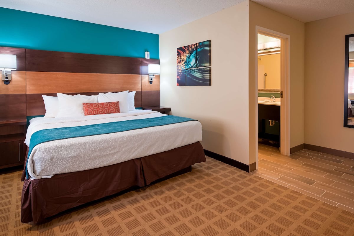 Habitación de hotel con cama, mesitas de noche, cabecera, un cuadro y baño visible a través de una puerta abierta.