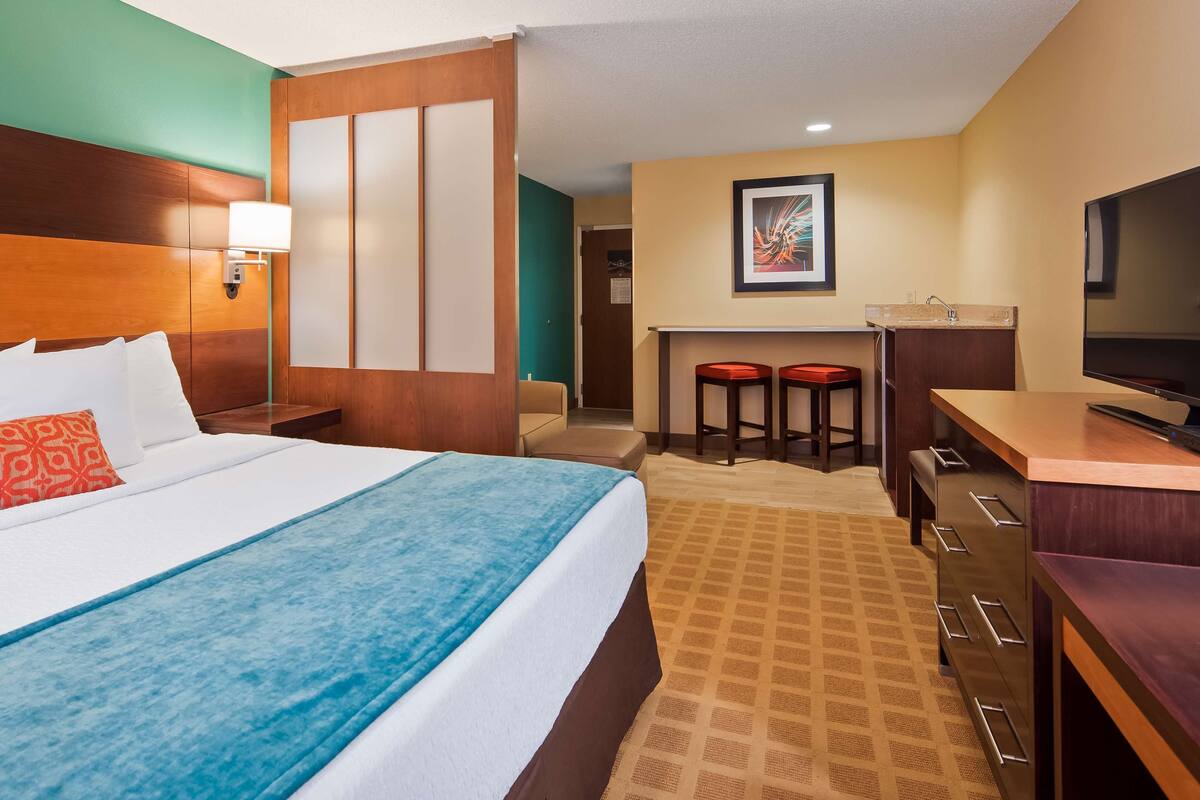 Una habitación de hotel con cama, escritorio, televisor y una pequeña kitchenette.