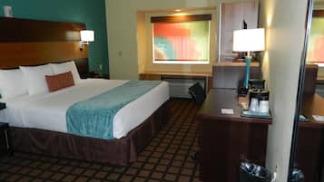 A hotel room with a bed, desk, and chair. There is a window with a view, a lamp, and a trash bin.