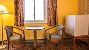 A room with two wooden chairs, a small round table, a white refrigerator, and a window with patterned curtains. - Rodeway Inn Lincoln I-93 (Lincoln)