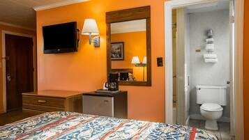 A hotel room with a bed, a TV, a desk with a coffee maker, a mirror, and a bathroom with a toilet and shower.