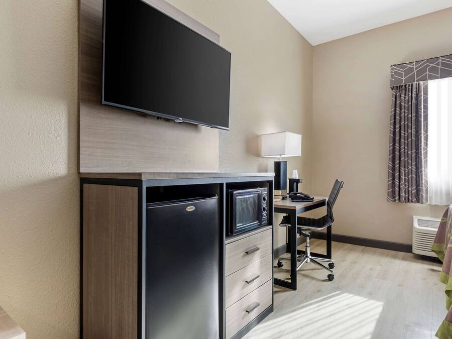 A hotel room with a flat-screen TV mounted on a wooden cabinet, a desk with a chair, a lamp, and a bed with a patterned bedspread.