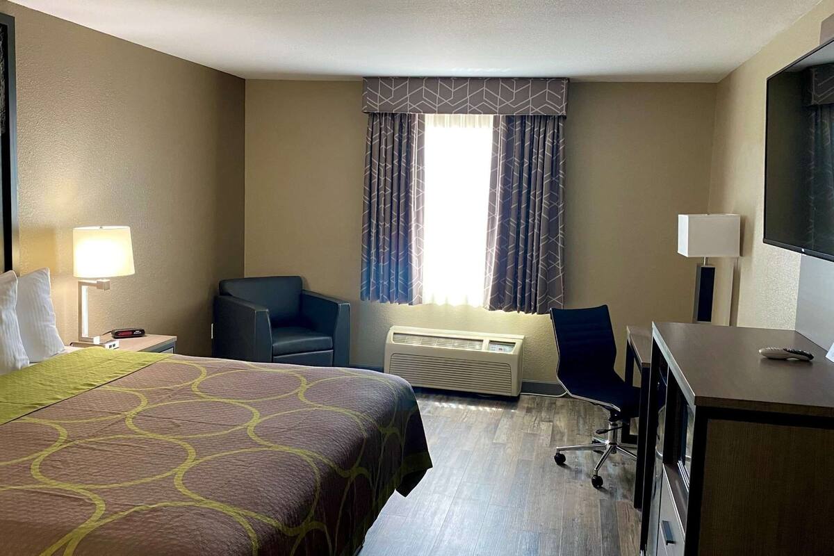 Quarto de hotel com cama, escrivaninha, cadeira, luminária, aparelho de ar condicionado e janela com cortinas.