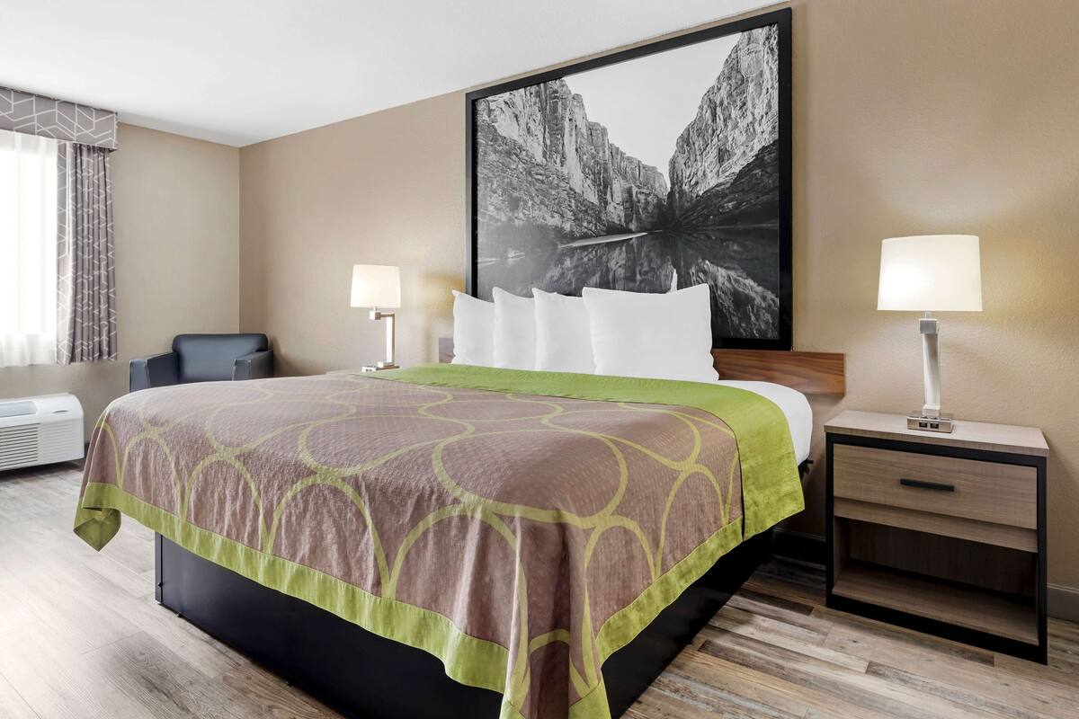 Quarto de hotel com uma cama grande, duas mesas de cabeceira com luminárias, um quadro grande na parede e uma janela com cortinas.