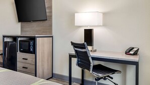 Ein Hotelzimmer mit Flachbildfernseher an der Wand, einem Schreibtisch mit Lampe, einem Sessel, einer Mikrowelle und einem Telefon.