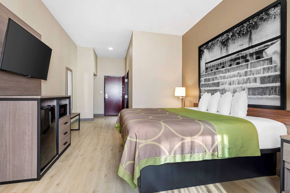 Quarto de hotel com cama, televisão na parede, escrivaninha e uma janela grande.