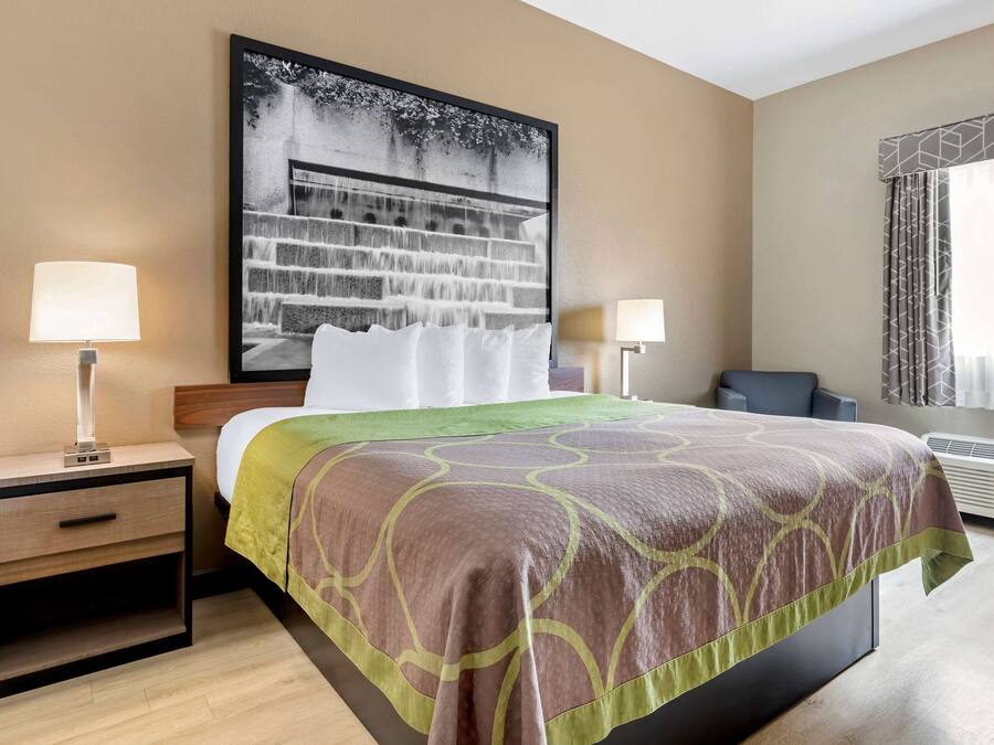 A hotel room with a bed, two bedside tables with lamps, a large framed picture on the wall, and an air conditioning unit.