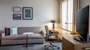 A hotel room with a sofa, a desk with a chair, a television, and framed pictures on the wall.