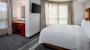 A hotel room with a bed, a TV mounted on the wall, a bathroom with a sink and mirror, and large windows with curtains.