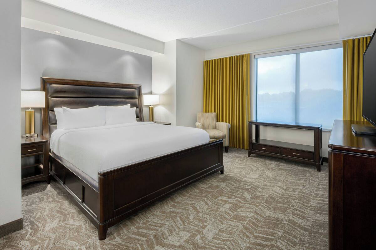A hotel room with a large bed, a TV, a chair, a desk, and a window with curtains.