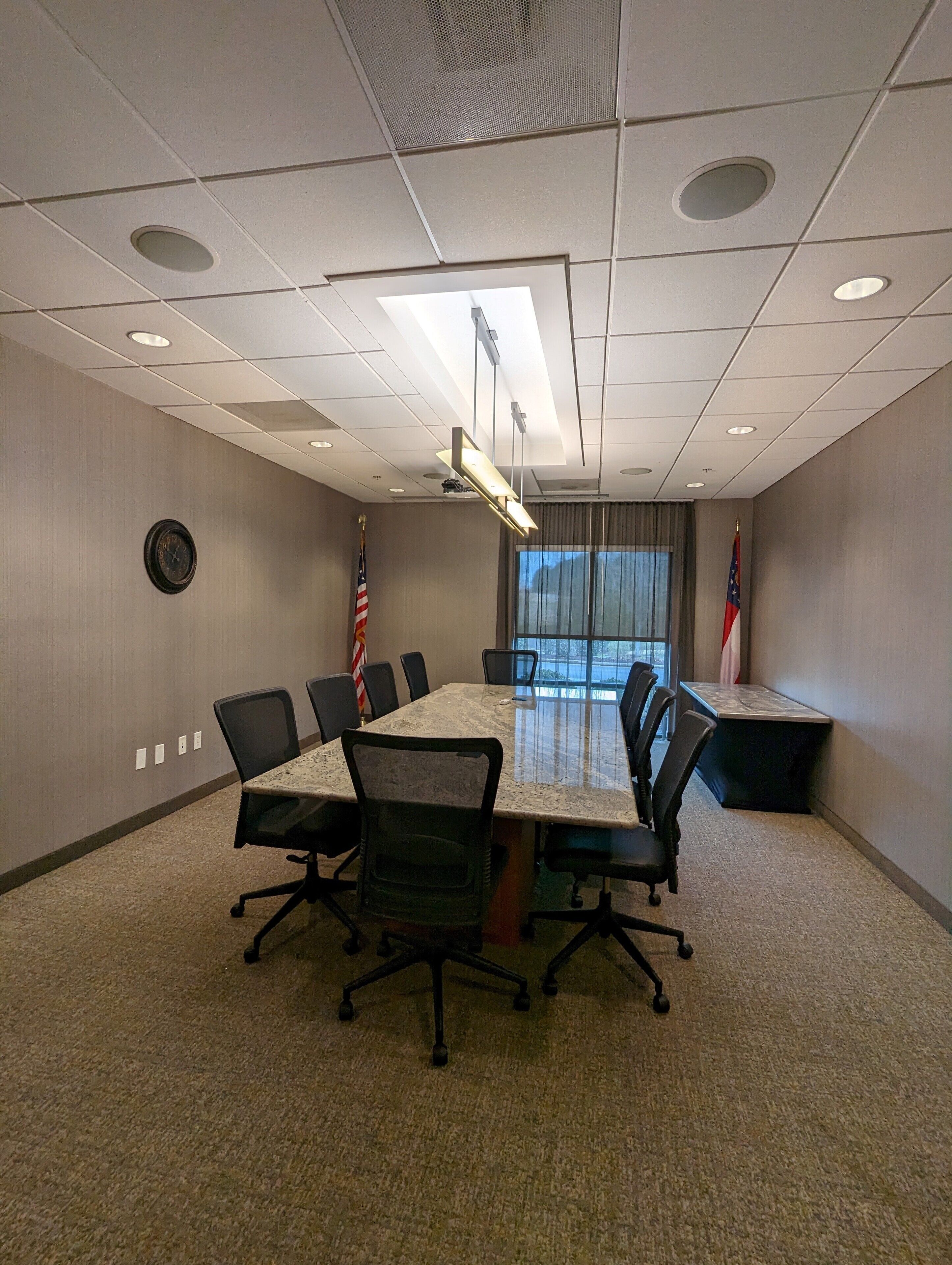 Uma sala de conferência com uma mesa comprida, cadeiras, bandeiras e um relógio na parede.