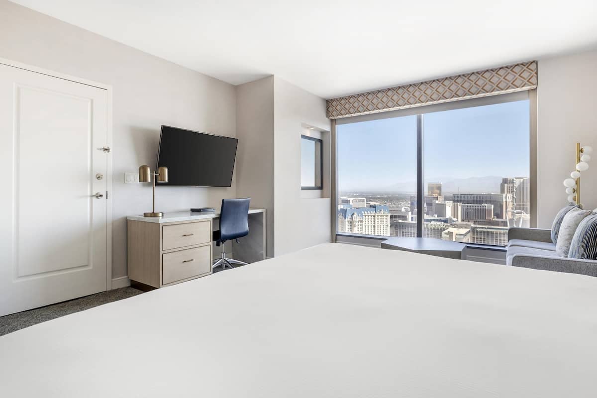 Hotelkamer met een groot bed, een bureau met stoel, een televisie en uitzicht op de stad door grote ramen.