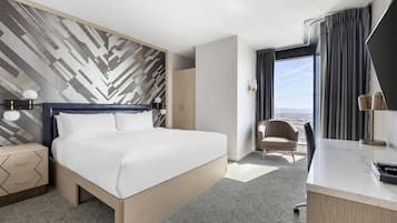 A modern hotel room with a large bed, a desk, a chair, and a view of the city.