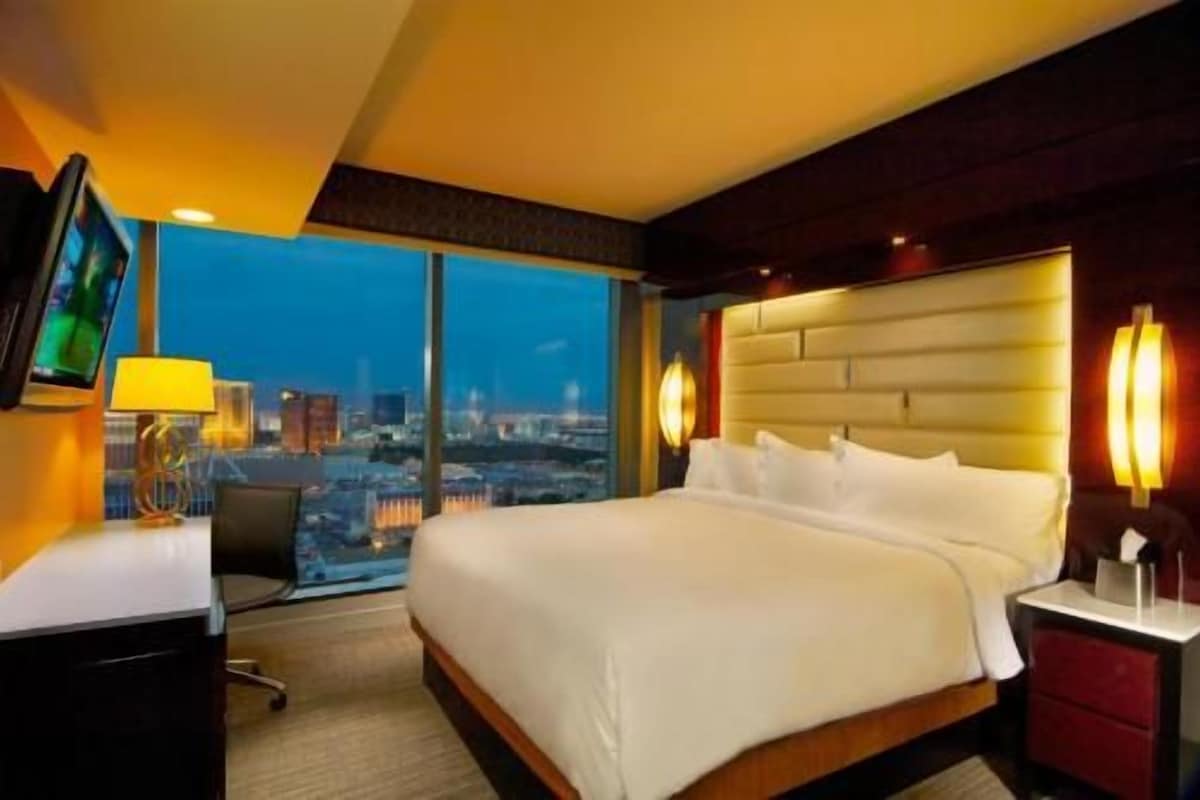 A hotel room with a large bed, a desk with a lamp, a TV, and a city view.