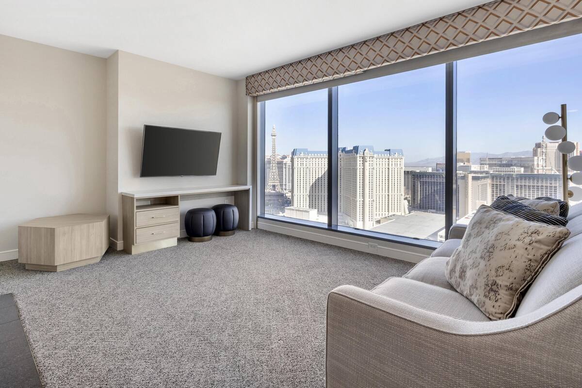 Een moderne hotelkamer met een groot raam met uitzicht op de stad, een flatscreen televisie aan de wand, een bank met een gekleurd kussen, en een klein houten bijzettafeltje.