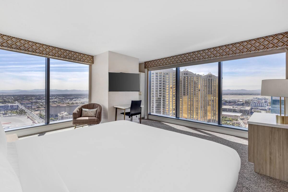 Een moderne hotelkamer met een groot bed, een bureau, een stoel en een televisie. De kamer heeft grote ramen met uitzicht op de stad.