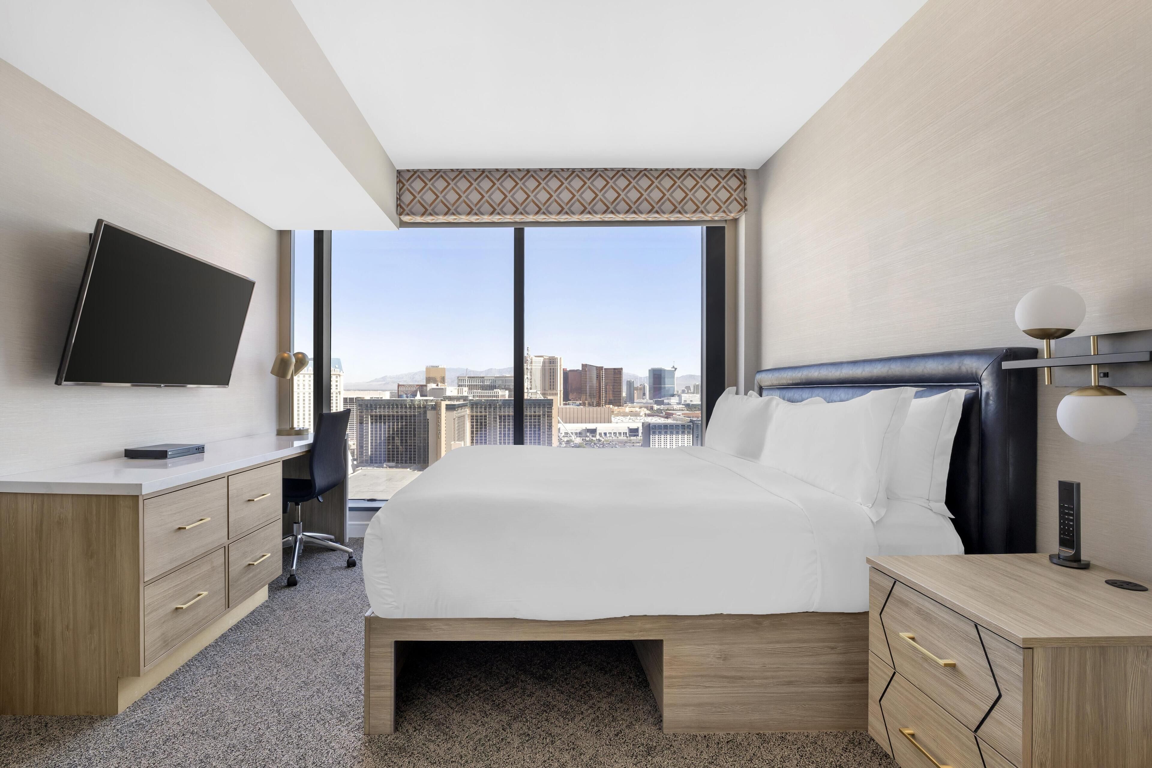 A hotel room with a large bed, a desk with a TV, and a city view.