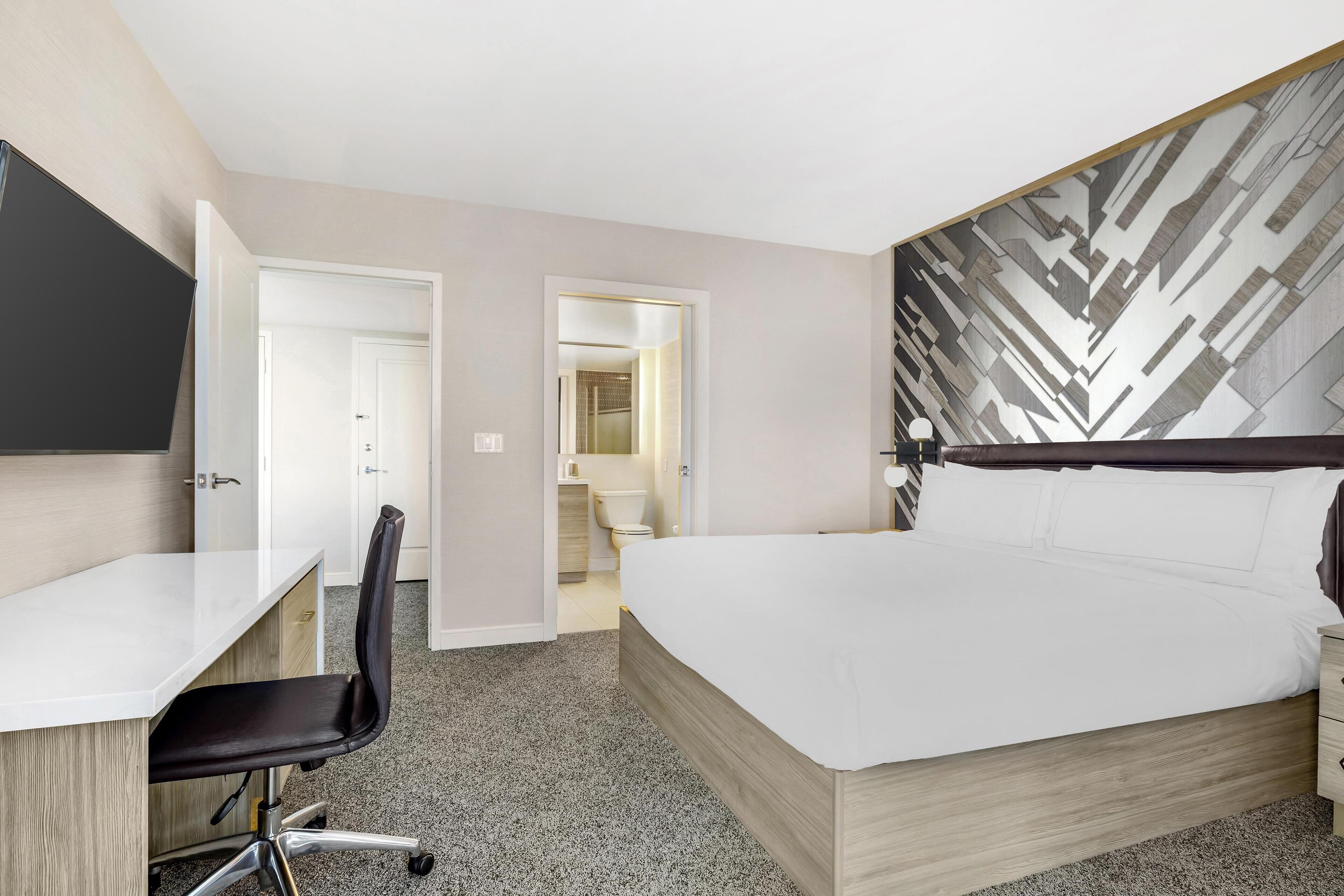 A hotel room with a bed, a desk, and a chair. There is a bathroom visible through an open door.