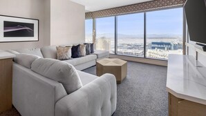A modern hotel room with a large window offering a city view, a grey sofa, a small coffee table, and a flat-screen TV.