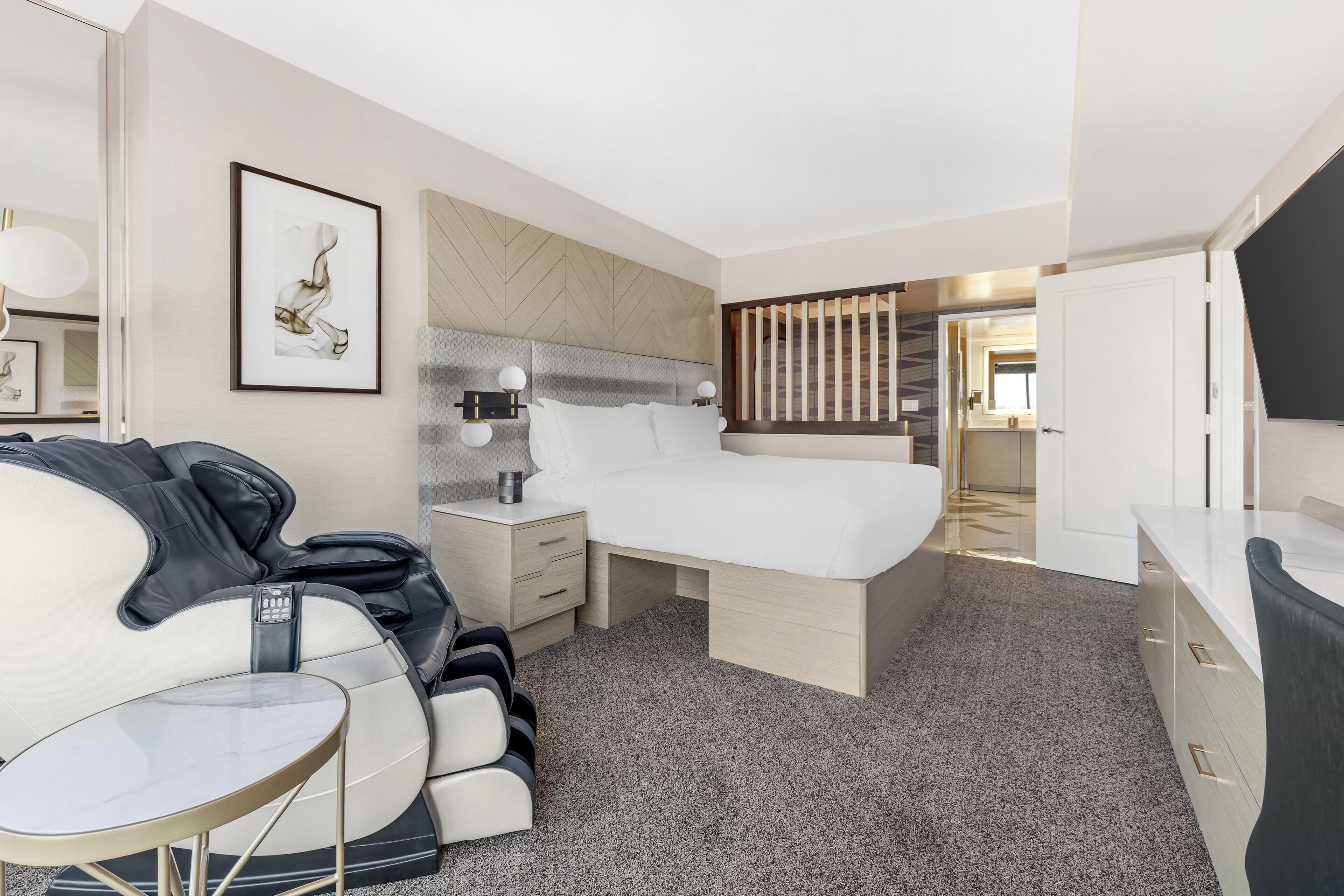 A modern hotel room with a bed, massage chair, bedside table, and a large mirror.
