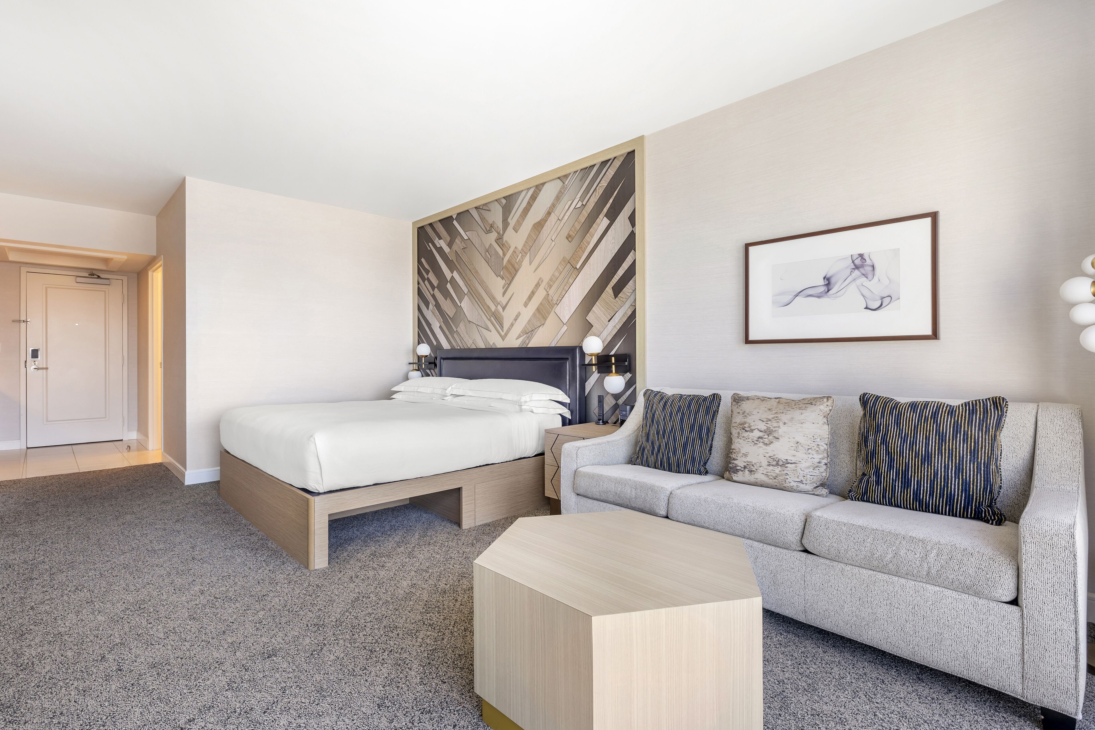 A modern hotel room with a bed, a sofa, a coffee table, and a wall with a geometric pattern.