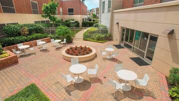 An outdoor patio area with round tables and white chairs, surrounded by brick walls and greenery.