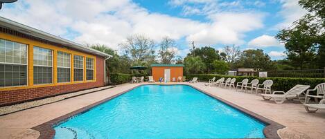 A rectangular swimming pool with a brick border, surrounded by a sandy area and a row of white lounge chairs.