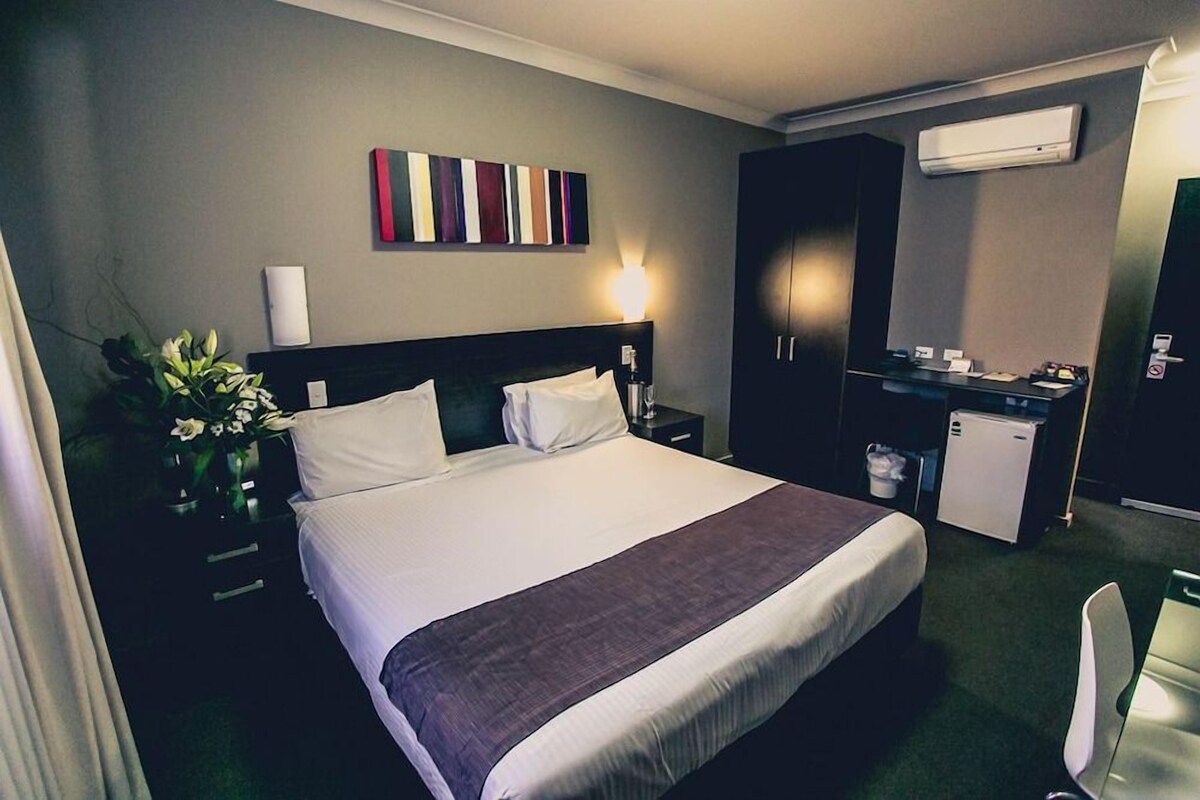 A hotel room with a bed, bedside table, lamp, air conditioning unit, and a small kitchenette.