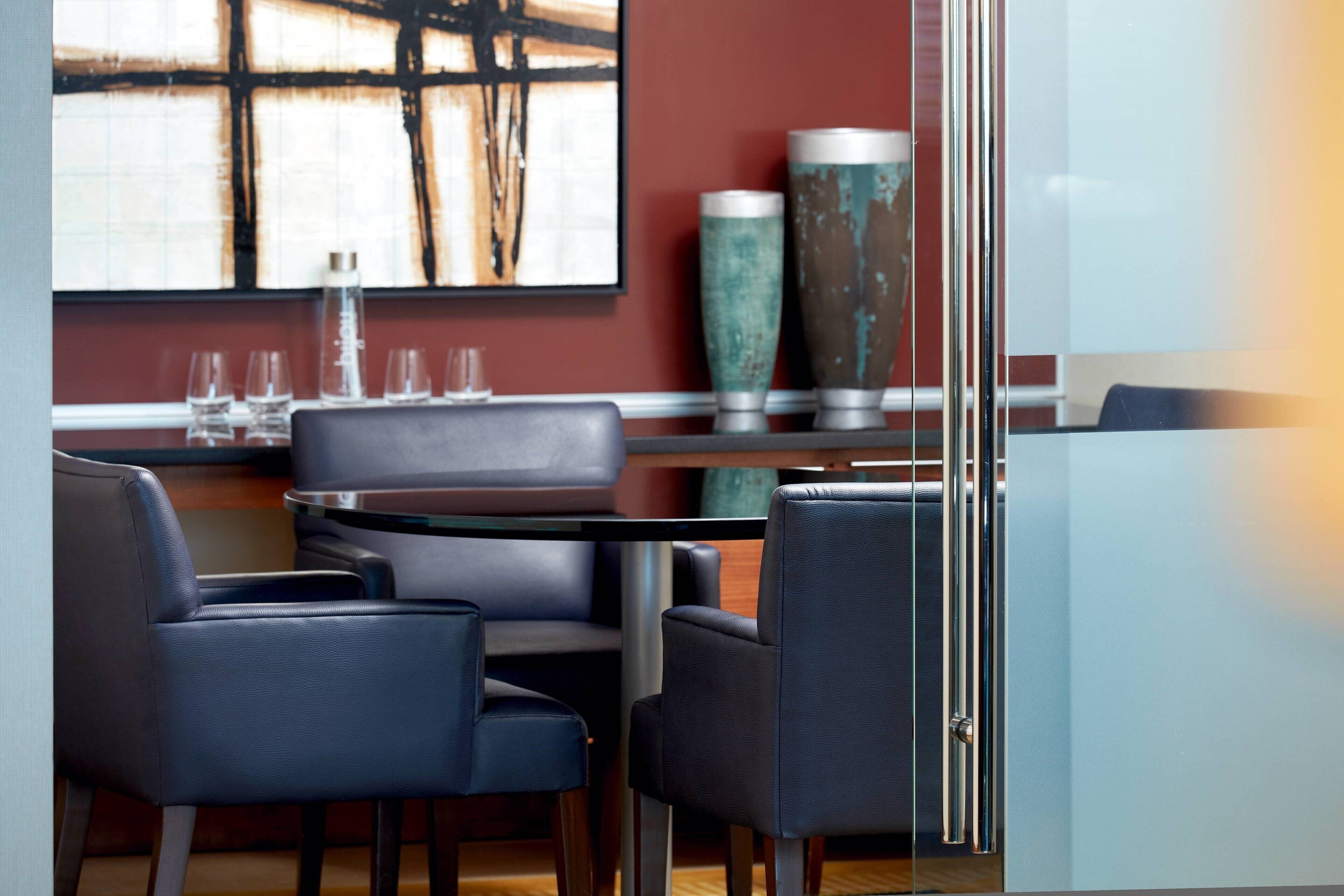 A modern dining area with a glass partition, leather chairs, a round table, and a large abstract painting on the wall.