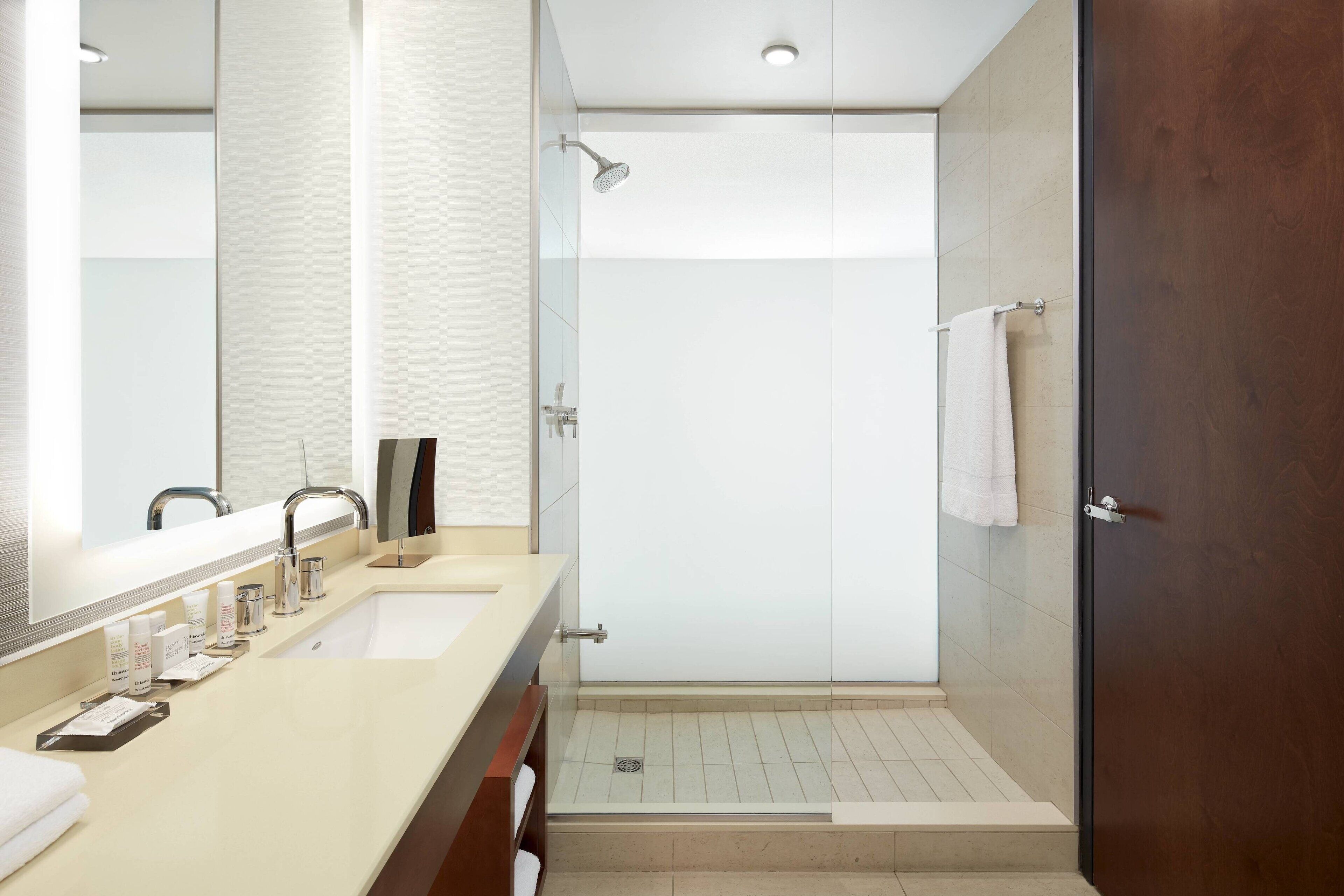 A modern bathroom with a double sink, a large mirror, a shower with a glass door, and a wooden door.