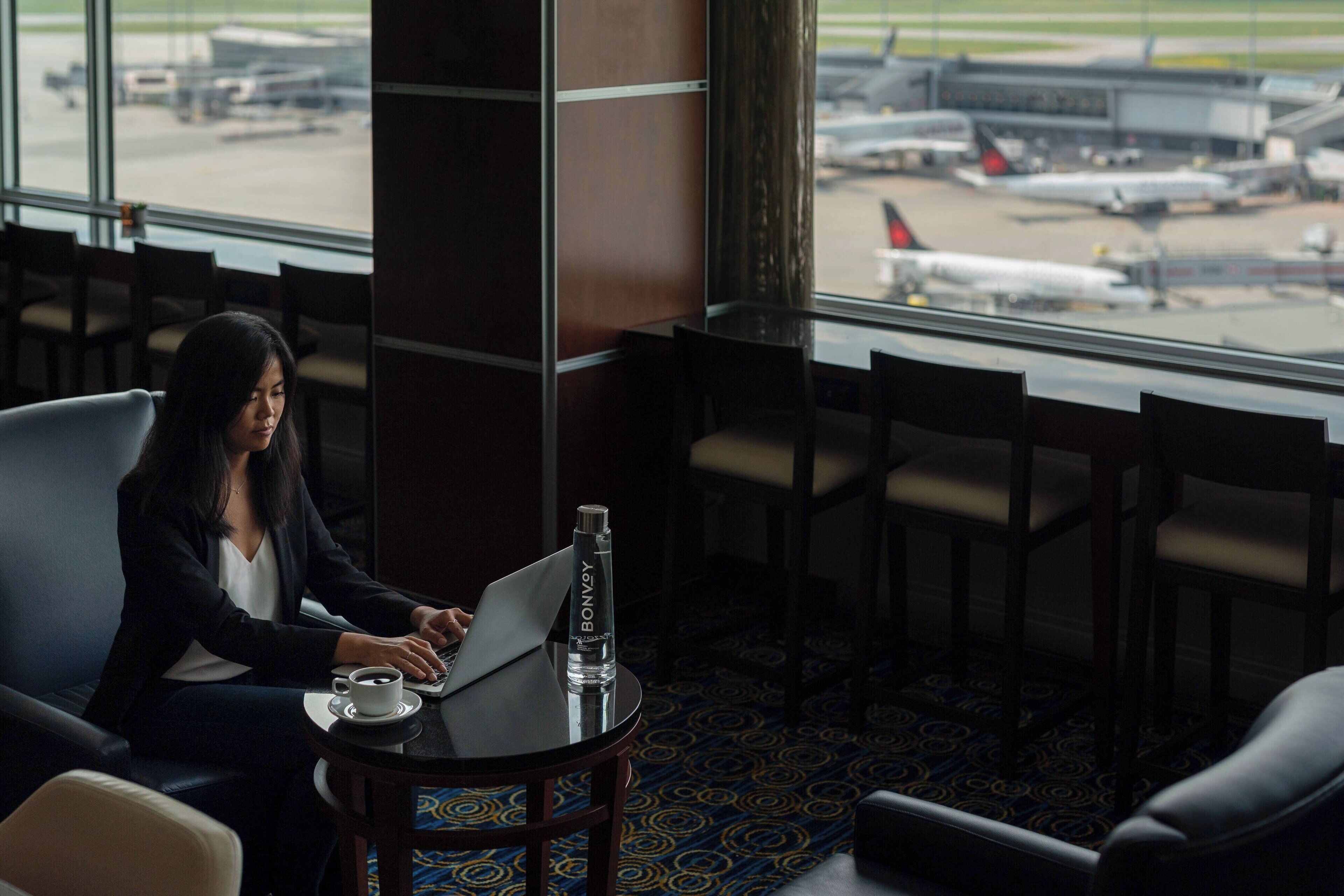 A woman working on a laptop in a hotel room with a view of the airport.