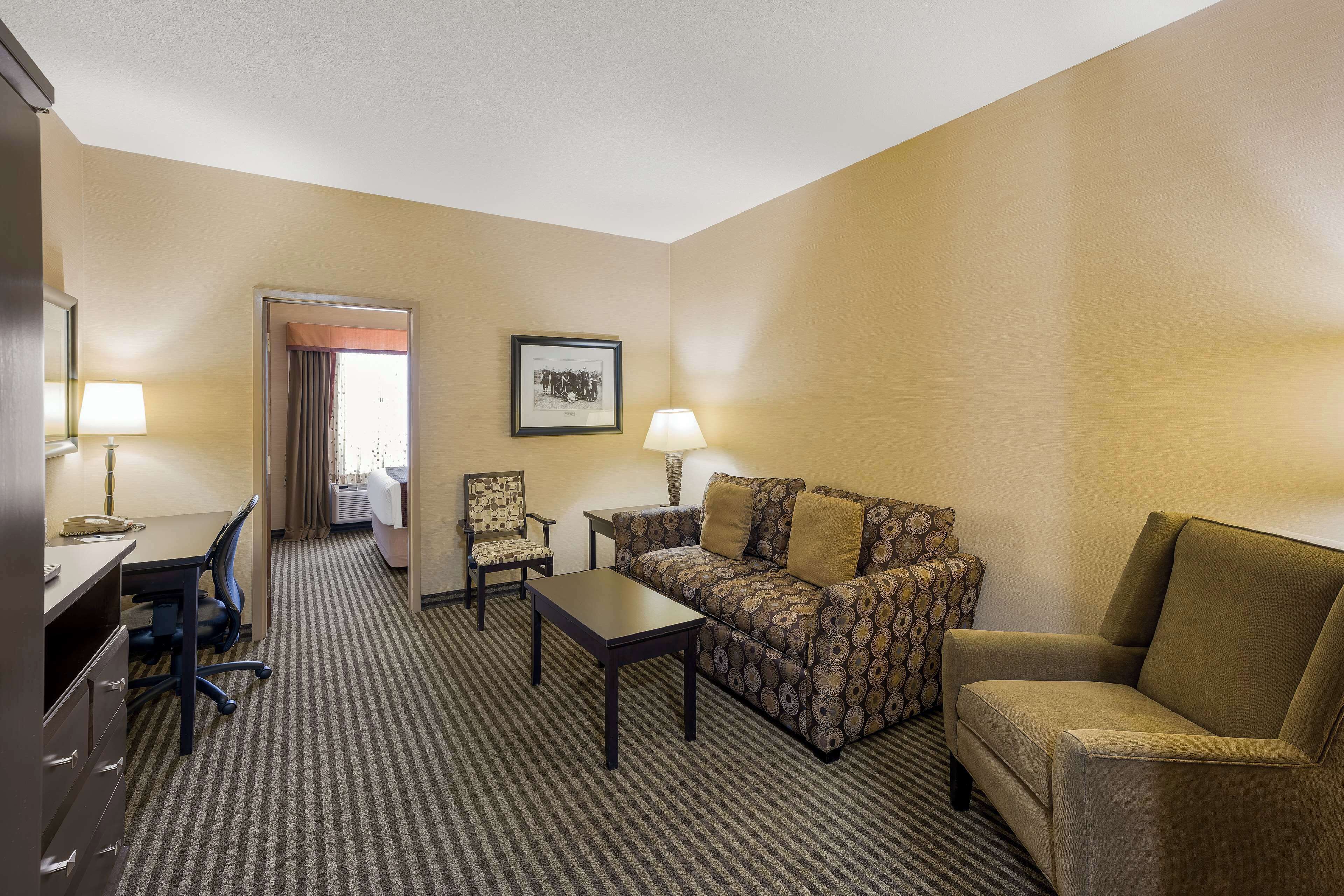 A hotel room with a sofa, armchair, desk, and chair. There is a framed picture on the wall and a lamp on the desk.