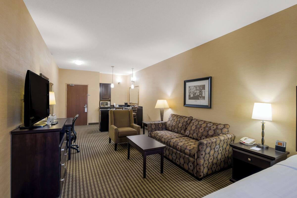 A hotel room with a bed, a desk with a computer, a sofa, a chair, a TV, and a lamp.