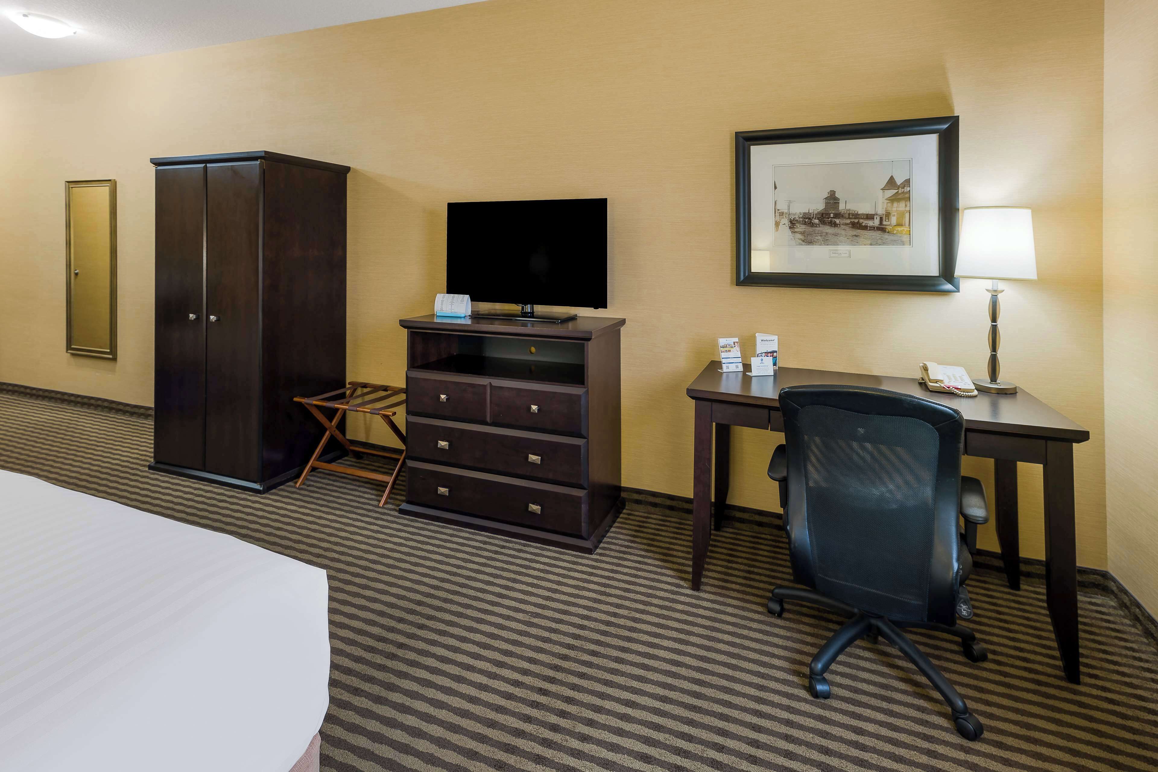 A hotel room with a bed, a desk with a chair, a television, a wardrobe, a small table with a lamp, and a framed picture on the wall.