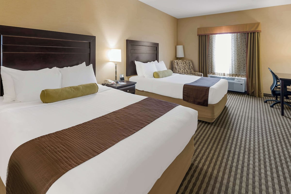A hotel room with two beds, a desk, a chair, a lamp, and an air conditioning unit.