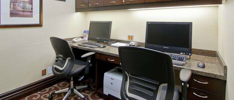 A hotel office with two desks, each with a computer, keyboard, and mouse. There are two office chairs, a printer, and a framed picture on the wall.