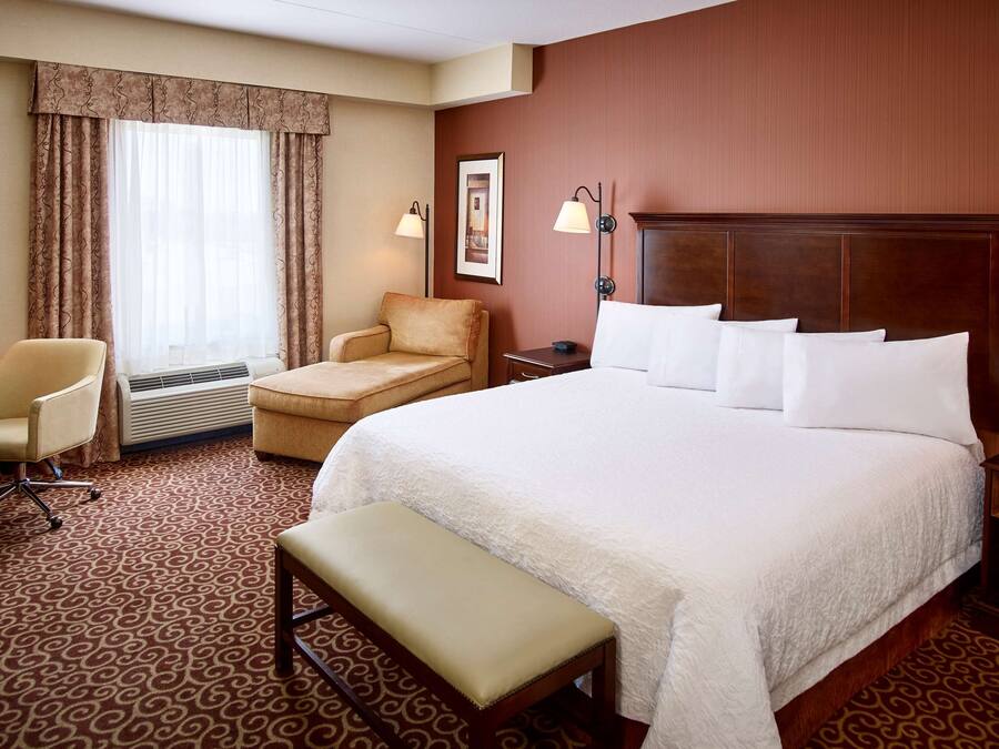 Quarto de hotel com uma cama grande, uma escrivaninha, uma cadeira, um banco e um quadro na parede.