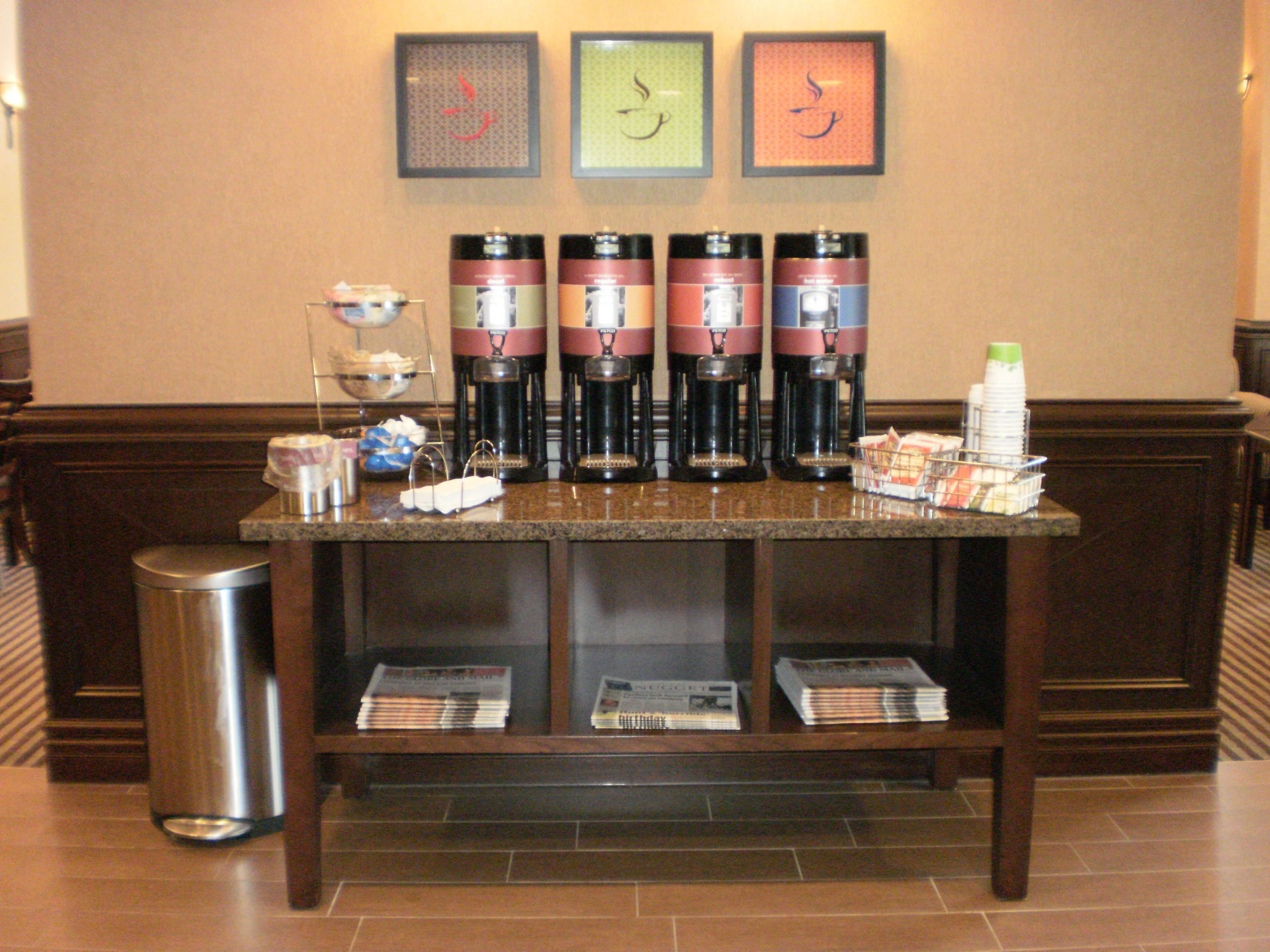 A coffee station with multiple coffee makers, a trash can, and magazines on a table.