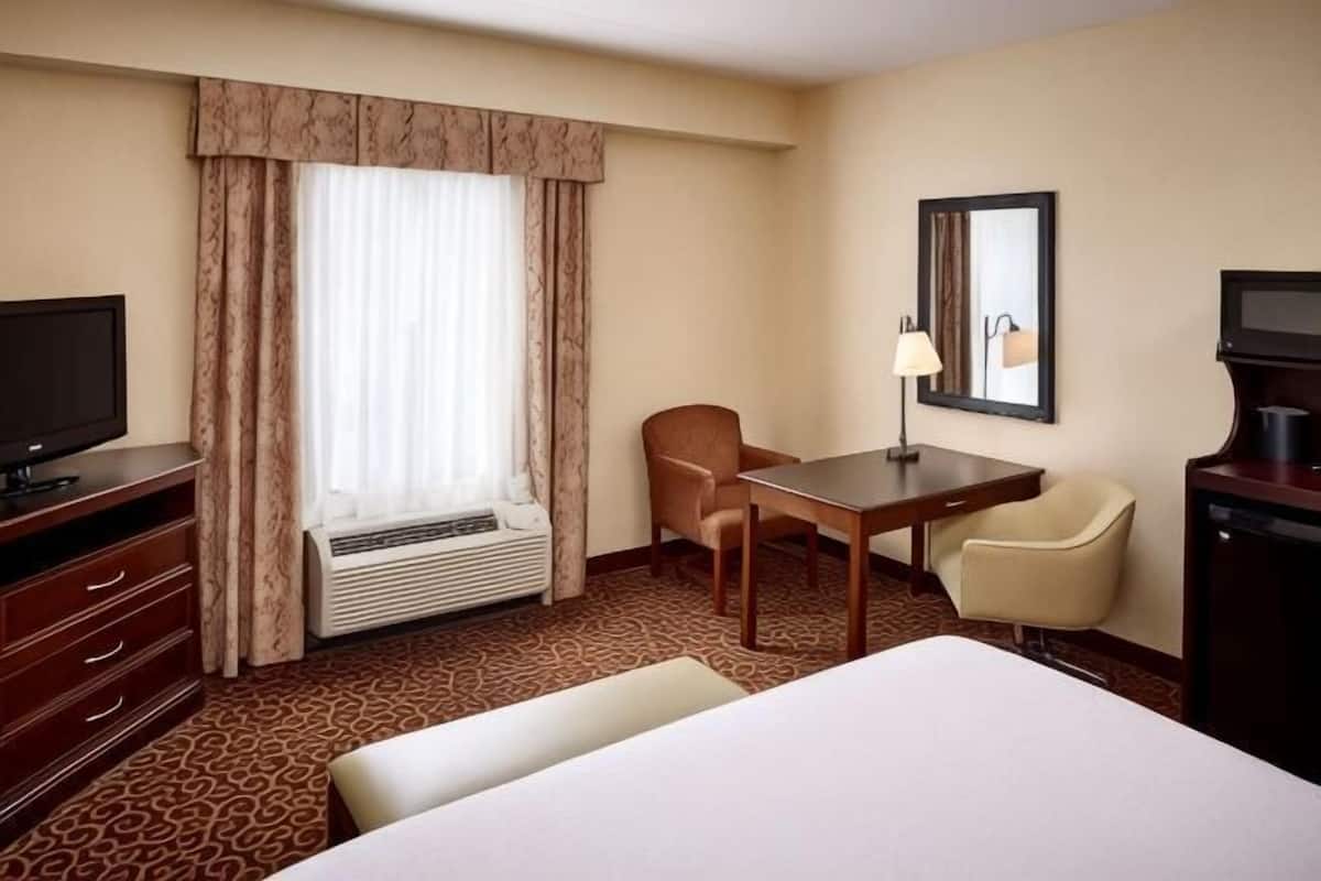 Una habitación de hotel con cama, escritorio, silla, televisor y ventana con cortinas.